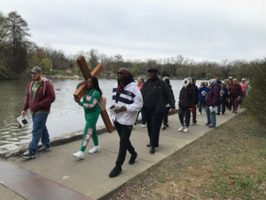 Picture of people walking beside a lake, one is carrying a cross